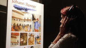 Sieviete ar austiņām klausās audio pie komiksu paneļa par Johann Renner hroniku