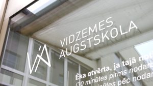 Tuvplānā redzams uzraksts "Vidzemes Augstskola" un tās logotips uz stikla durvīm vai loga.