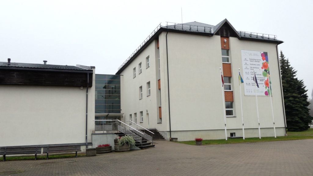 Balta, moderna Vidzemes Augstskolas ēka ar stiklotu pāreju, afišu un karogu mastiem priekšā.