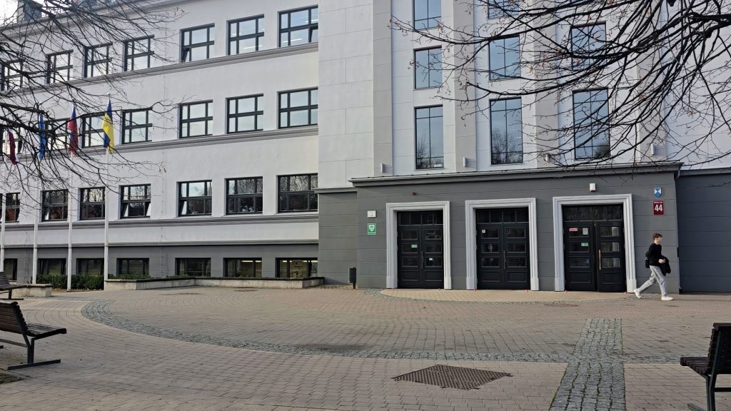 Galvenā ieeja Jelgavas Valsts ģimnāzijas ēkā ar trim tumšām durvīm un bruģētu laukumu.