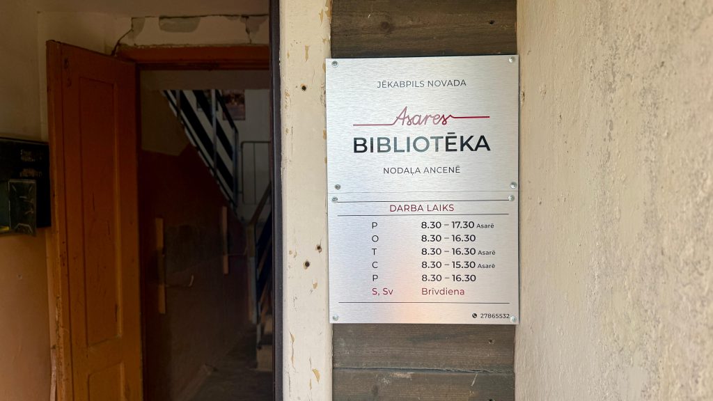 Plāksne ar Asares bibliotēkas Ancenes nodaļas nosaukumu un darba laiku uz vecas ēkas fasādes