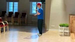 Elviss Strazdiņš ar mikrofonu vada nodarbību vai lekciju zālē pie skatuves.