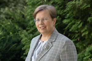VVAAD Augu karantīnas departamenta direktore Gunita Šķupele smaidot skatās kamerā, tērpusies pelēkā rūtainā žaketē uz zaļu koku fona