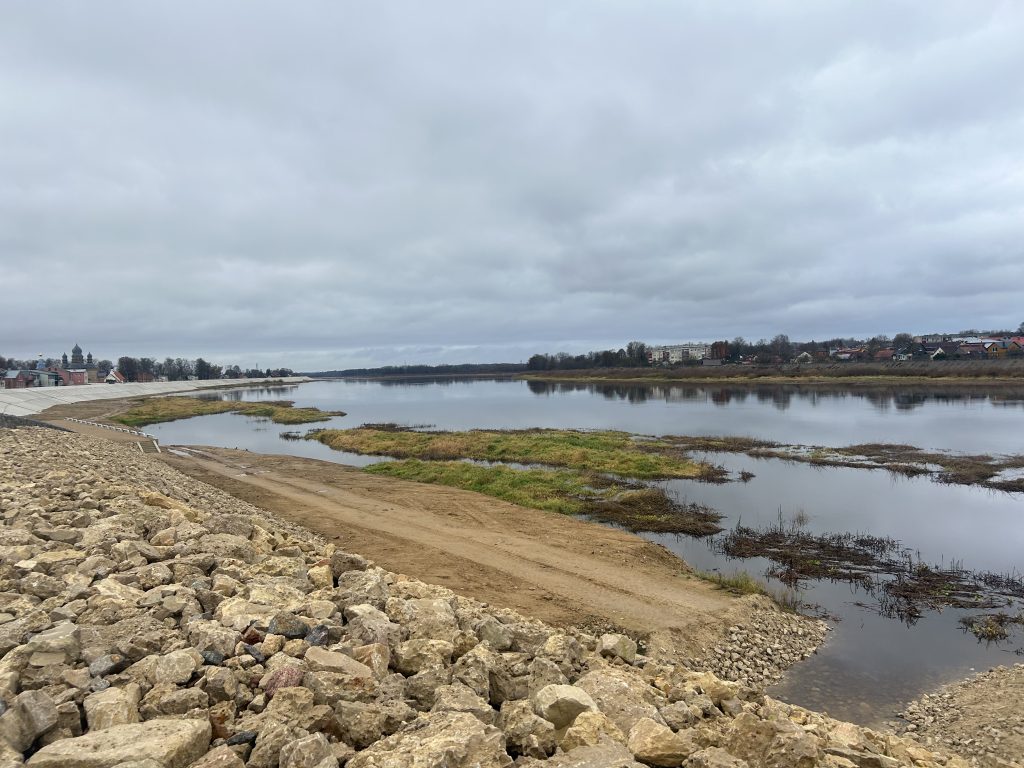 Plašs skats uz atjaunoto Daugavas aizsargdambi, ko veido lieli akmeņu krāvumi, ar mierīgu upes tecējumu un Jēkabpils pilsētu fonā