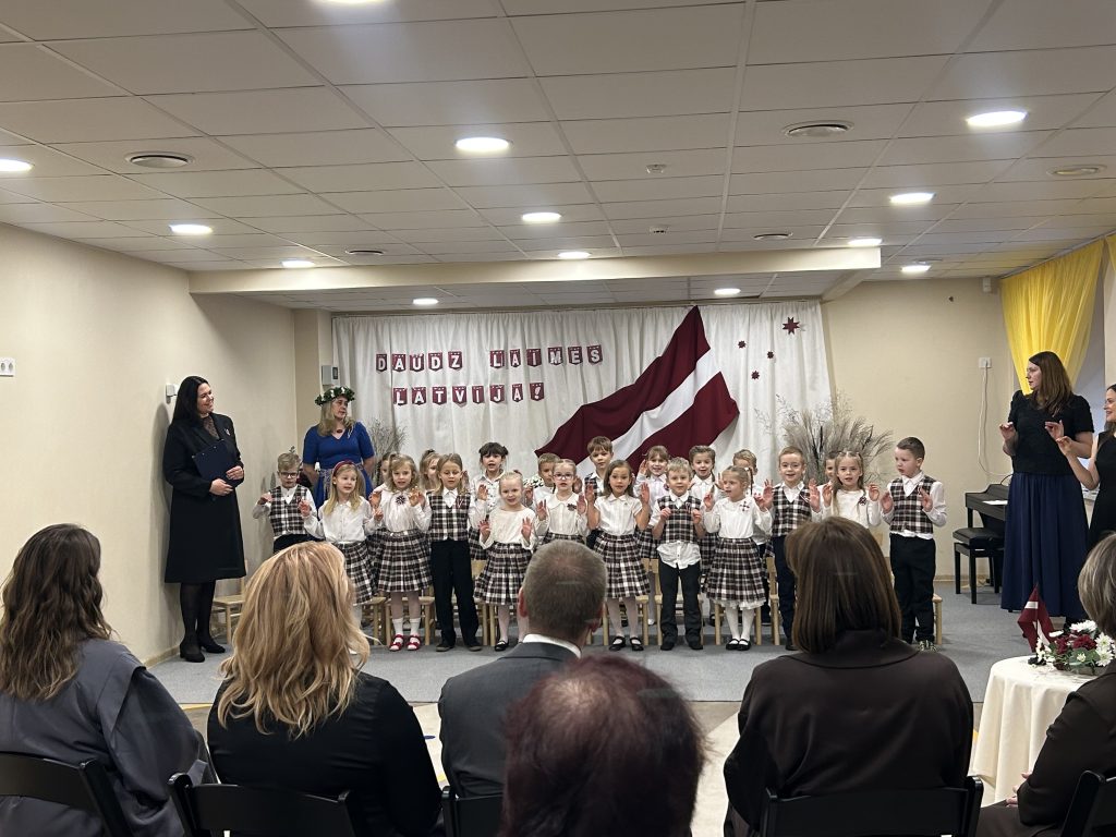 Bērni stāv rindā uz skatuves ar paceltām rokām, dziedot vai runājot dzejoli.