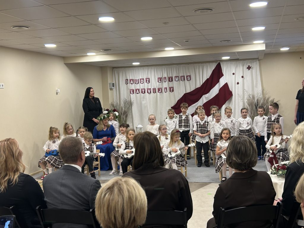 Bērnu grupa tautas tērpu stila apģērbā stāv uz skatuves, turot dekoratīvas ģitāras, uzstājoties Valsts prezidentam un skatītājiem.