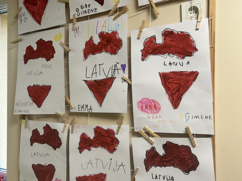 Bērnu zīmējumi, kas attēlo Latvijas karti un karogu krāsas, ar piestiprinātiem veļas knaģiem pie sienas.