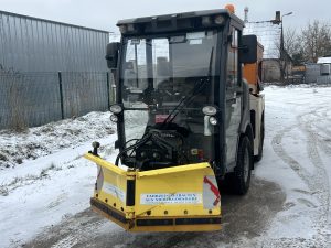 Kompakta sniega tīrīšanas mašīna ar dzeltenu priekšējo lāpstu stāv sniegotā industriālā teritorijā.