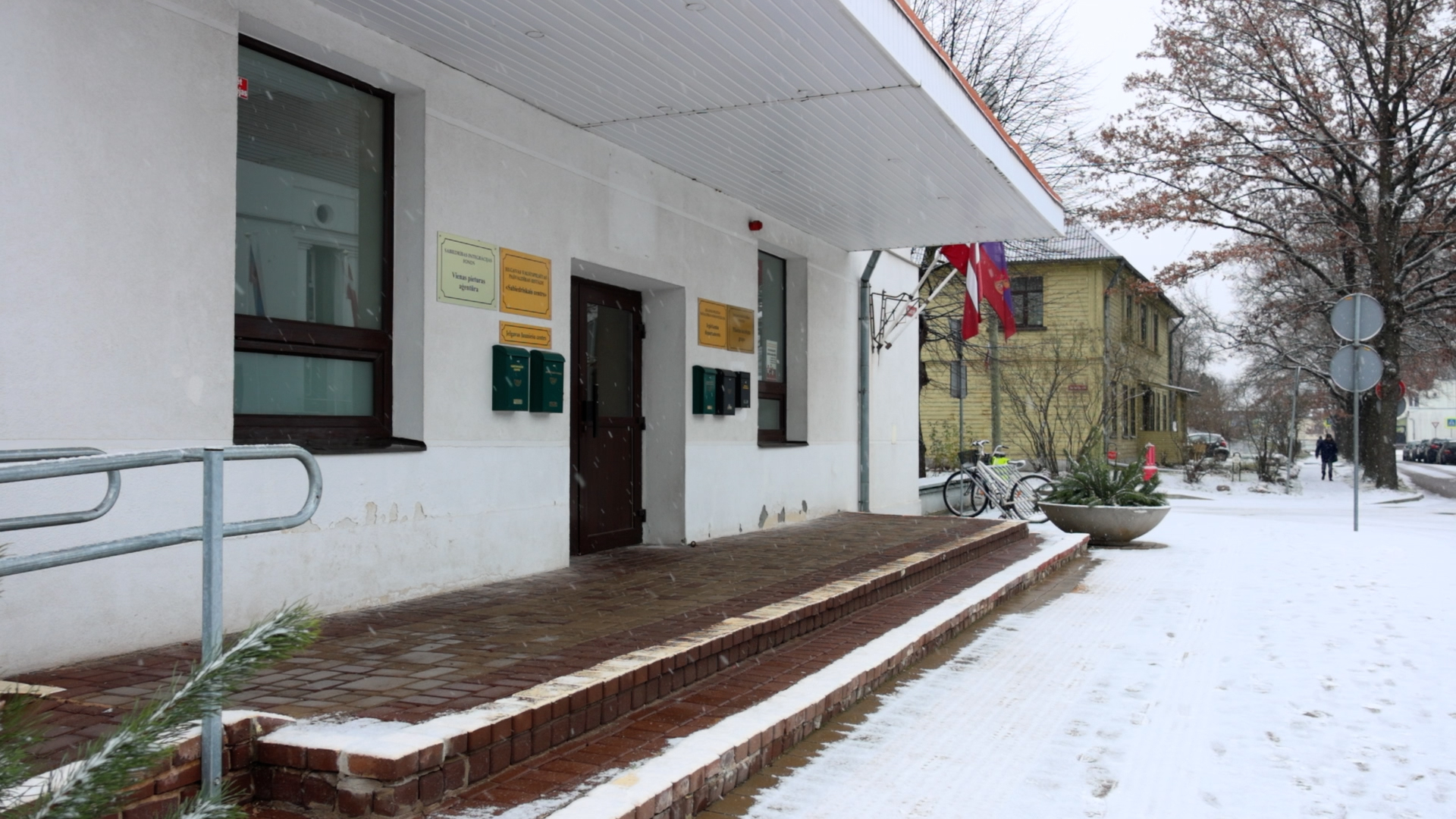 Jelgavas Sabiedriskā centra ēka ziemā ar valsts karogiem un informācijas plāksnēm pie ieejas.