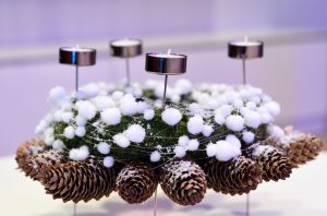 Adventes vainags, kas veidots no sūnu pamatnes, dekorēts ar baltām sniega bumbiņām un četriem metāla svecīšu turētājiem, ko ieskauj egļu čiekuri.