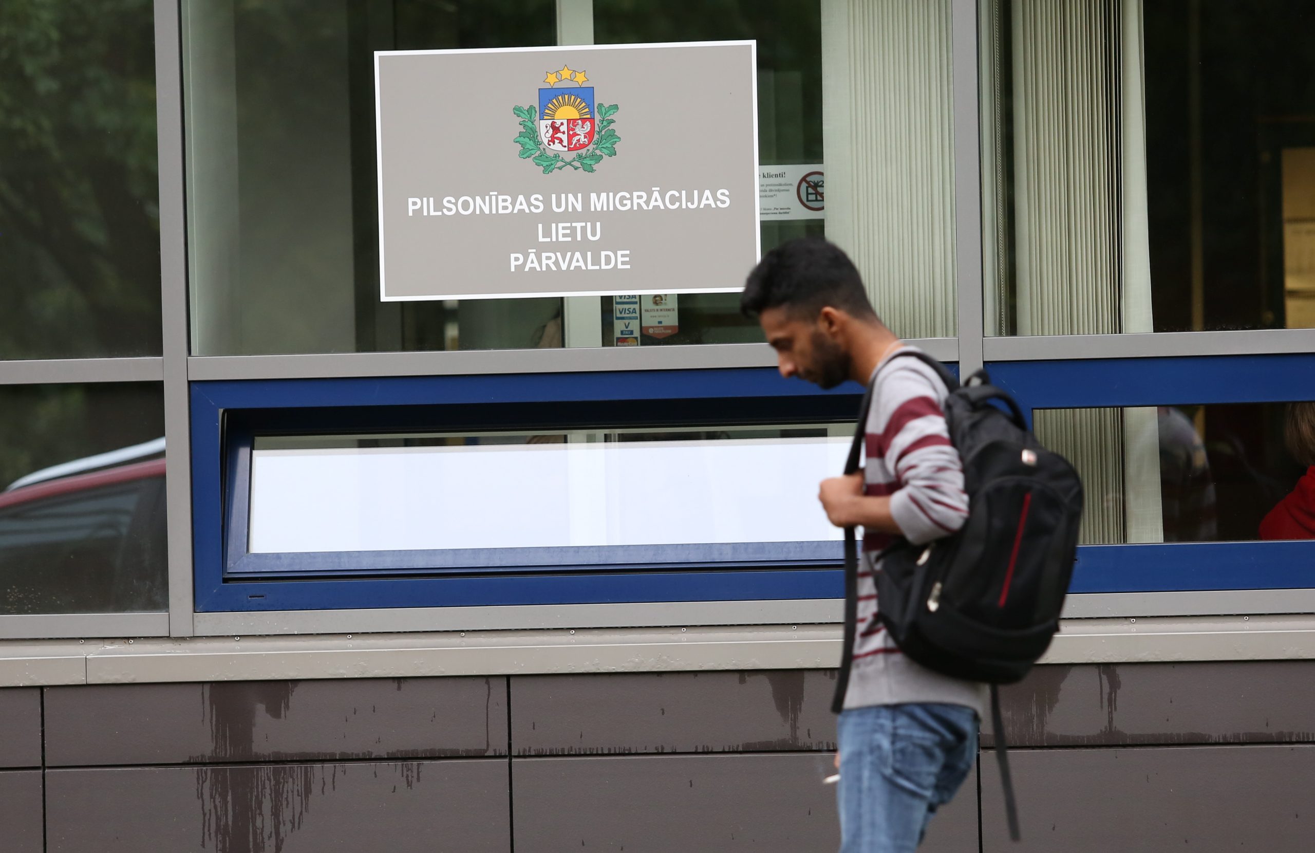 Cilvēks ar mugursomu iet garām ēkai, virs kuras logiem redzama Pilsonības un migrācijas lietu pārvaldes (PMLP) zīme.