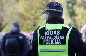 Aizmugures skats uz Rīgas pašvaldības policijas darbinieku gaiši zaļā (neona) vestē ar uzrakstu "RĪGAS PAŠVALDĪBAS POLICIJA", stāvot pūlī vai pasākumā.