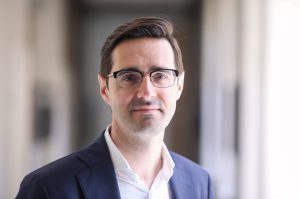 Tuvplāna portrets, kurā redzams vīrietis ar brillēm, Mārtiņš Lazdovskis, uzvalka žaketē un baltā kreklā.