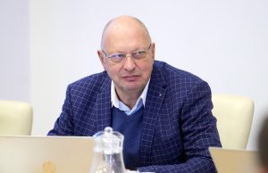 Jānis Lediņš sēž pie galda sanāksmē, skatoties uz priekšu, ģērbies žaketē ar rūtiņu rakstu.