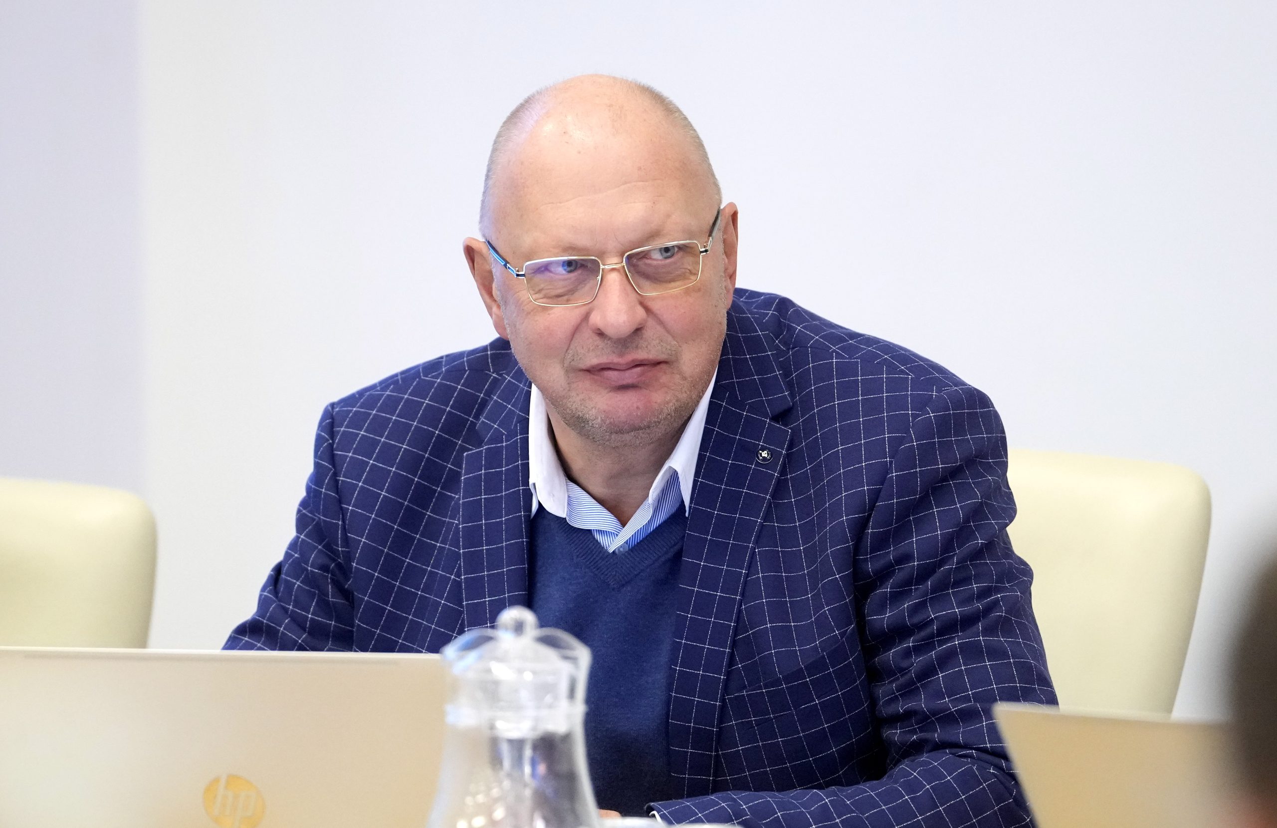 Jānis Lediņš sēž pie galda sanāksmē, skatoties uz priekšu, ģērbies žaketē ar rūtiņu rakstu.