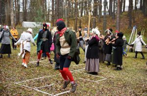 Masku gājiens Brīvdabas muzejā Mārtiņdienā, kurā tēli ar maskām dejo un lec laukumā, kamēr apkārt stāv pūlis un muzikanti.