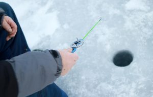 Tuvplāns, kurā redzama vīrieša roka, kas tur mazu, zilu ziemas makšķeri virs ledū izurbtā āliņģa.