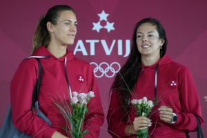 Divas sportistes Latvijas sporta formas tērpos ar ziediem rokās, pozējot uz sarkanā fona ar Latvijas olimpiskajiem simboliem.
