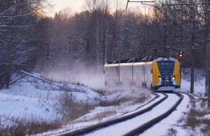 Dzeltenais un pelēkais elektrovilciens "vivi" ziemā pārvietojas pa sliedēm, atstājot aiz sevis sniega putekļus.