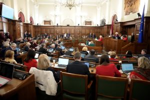 Saeimas sēdes zāle sēdes laikā, deputāti sēž pie galdiem ar datoriem, priekšplānā redzami Latvijas un Eiropas Savienības karogi.