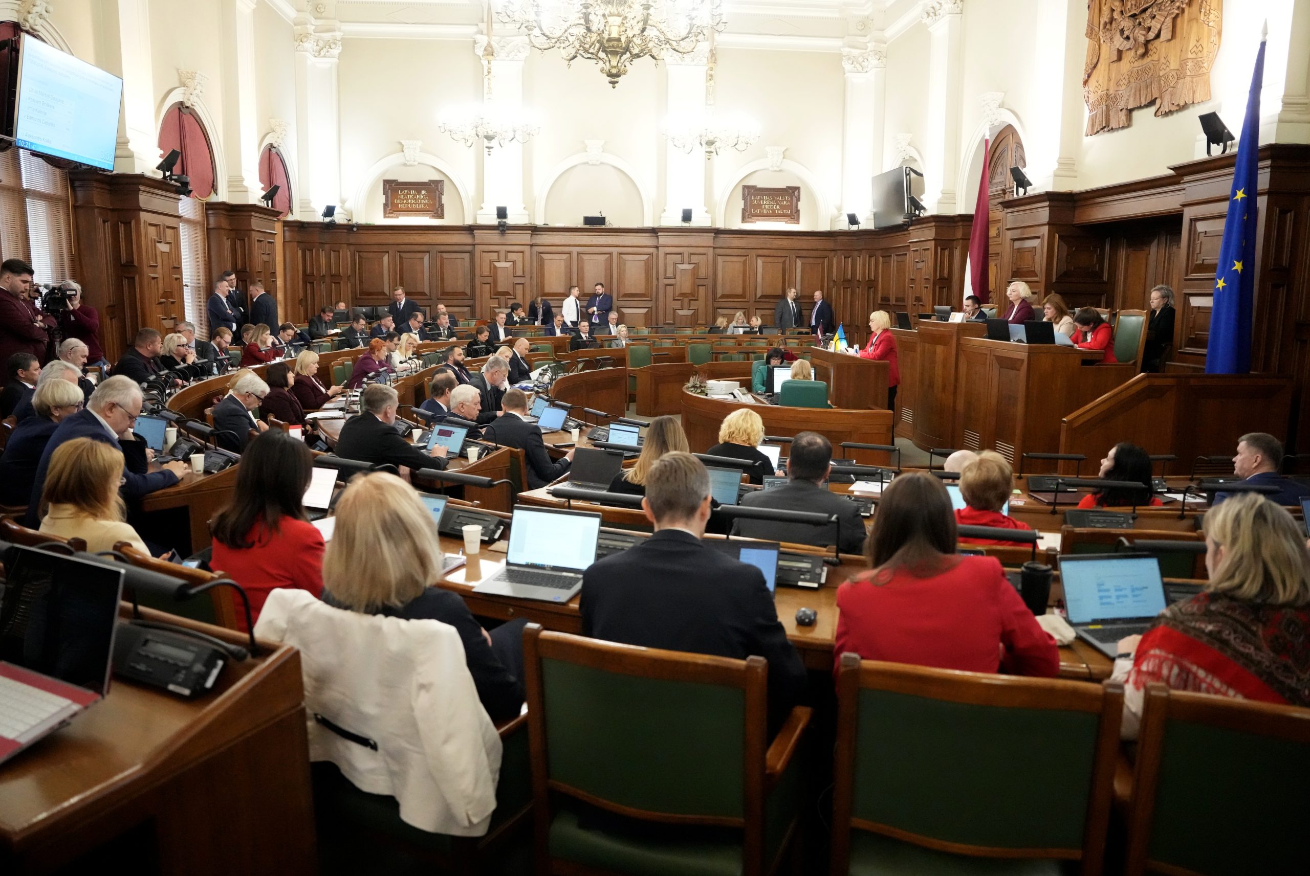 Saeimas sēdes zāle sēdes laikā, deputāti sēž pie galdiem ar datoriem, priekšplānā redzami Latvijas un Eiropas Savienības karogi.