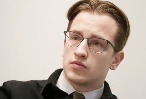 Jauns vīrietis ar brillēm un nopietnu sejas izteiksmi, skatoties sāņus biroja vidē.