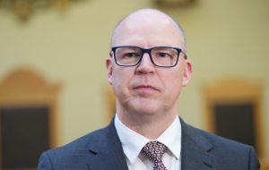 Tuvplāna portrets uzvalkā tērptam vīrietim ar brillēm.