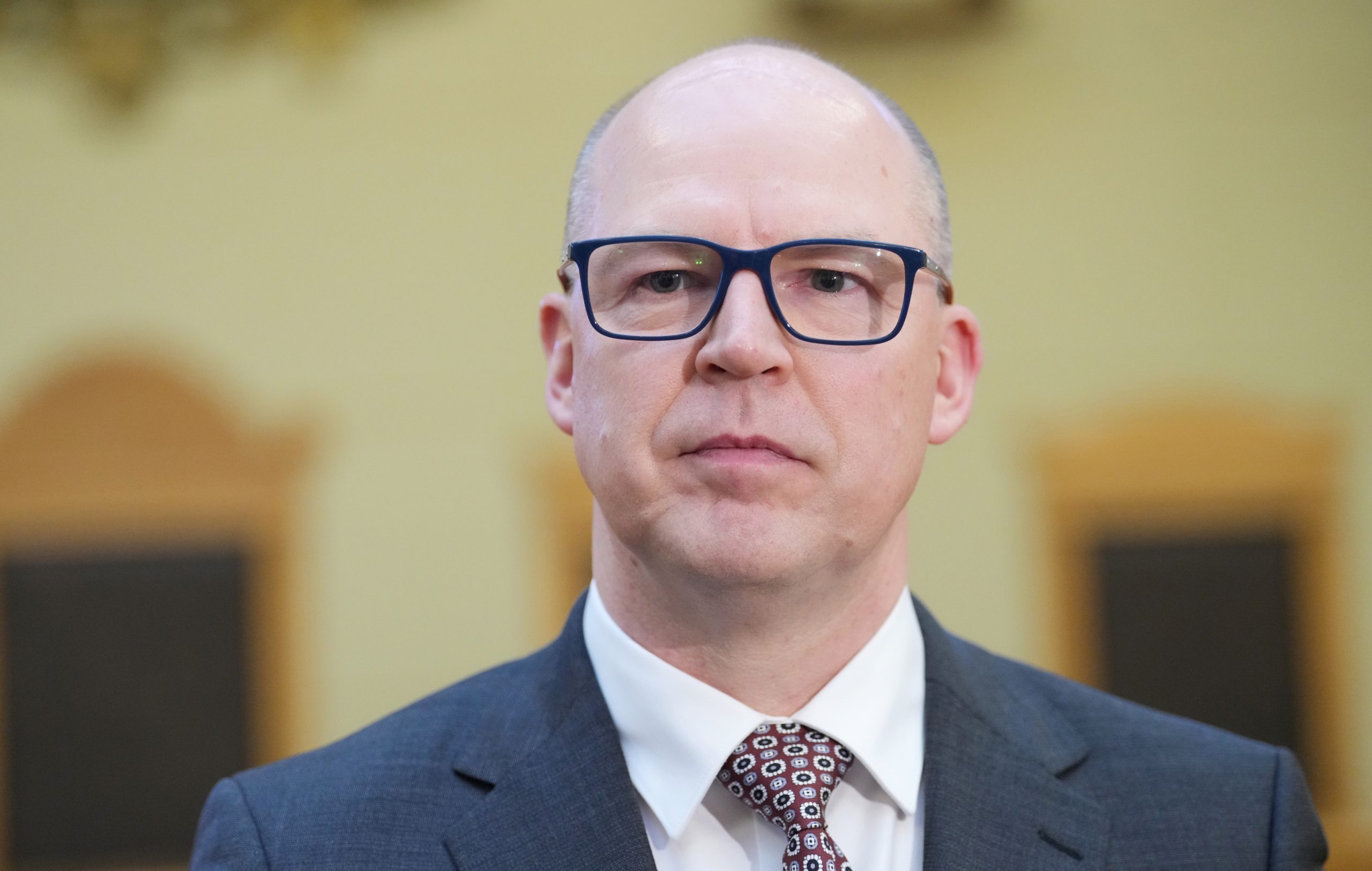Tuvplāna portrets uzvalkā tērptam vīrietim ar brillēm.