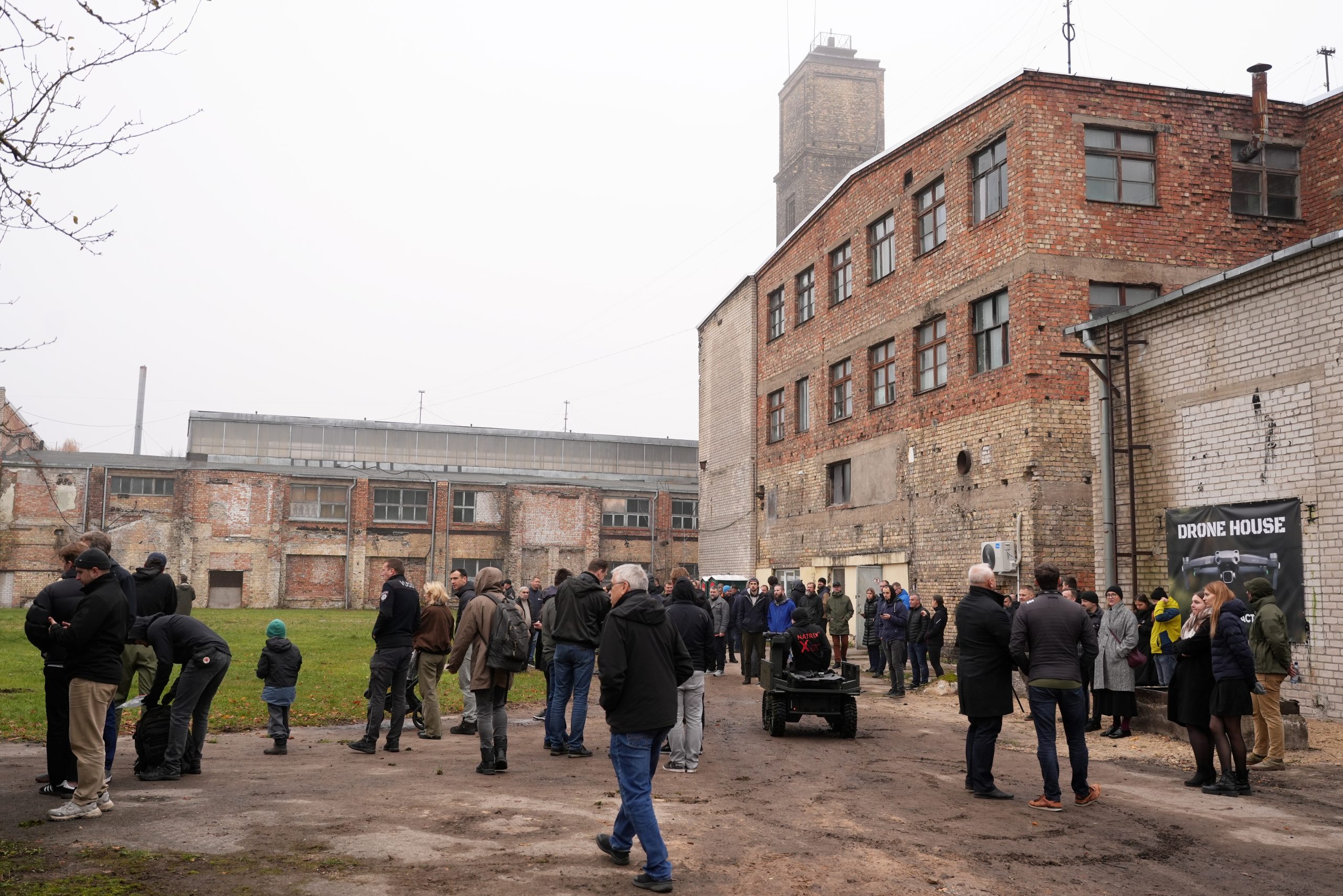 Cilvēku grupa pulcējusies dubļainā laukumā pie vecas ķieģeļu ēkas, atklājot dronu mācību centru 