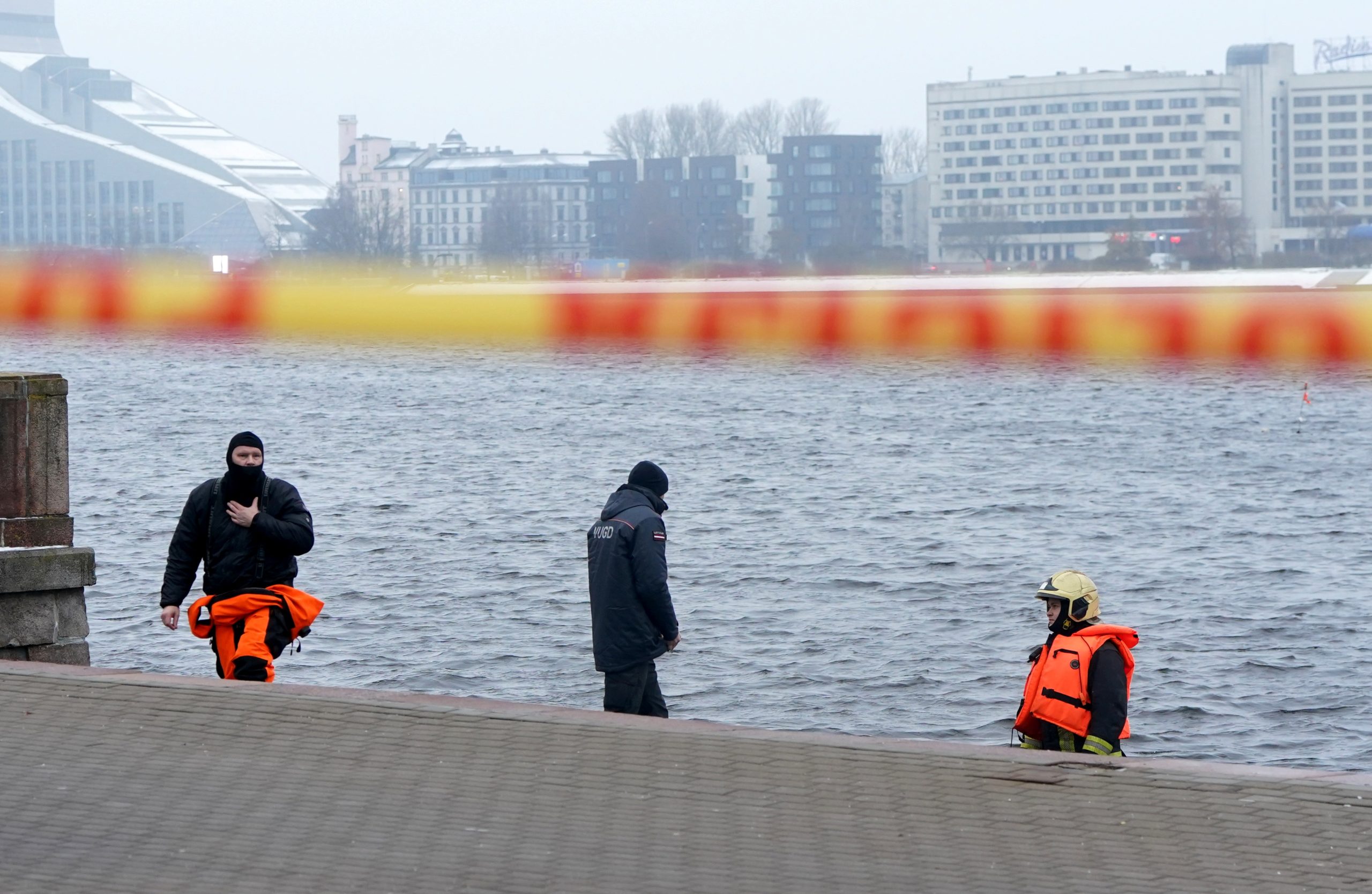 Glābēji un policijas darbinieki Daugavas krastmalā, kur norobežota automašīnas izcelšanas darbu vieta. Fonā redzama Rīgas ainava, ieskaitot Gaismas pili.