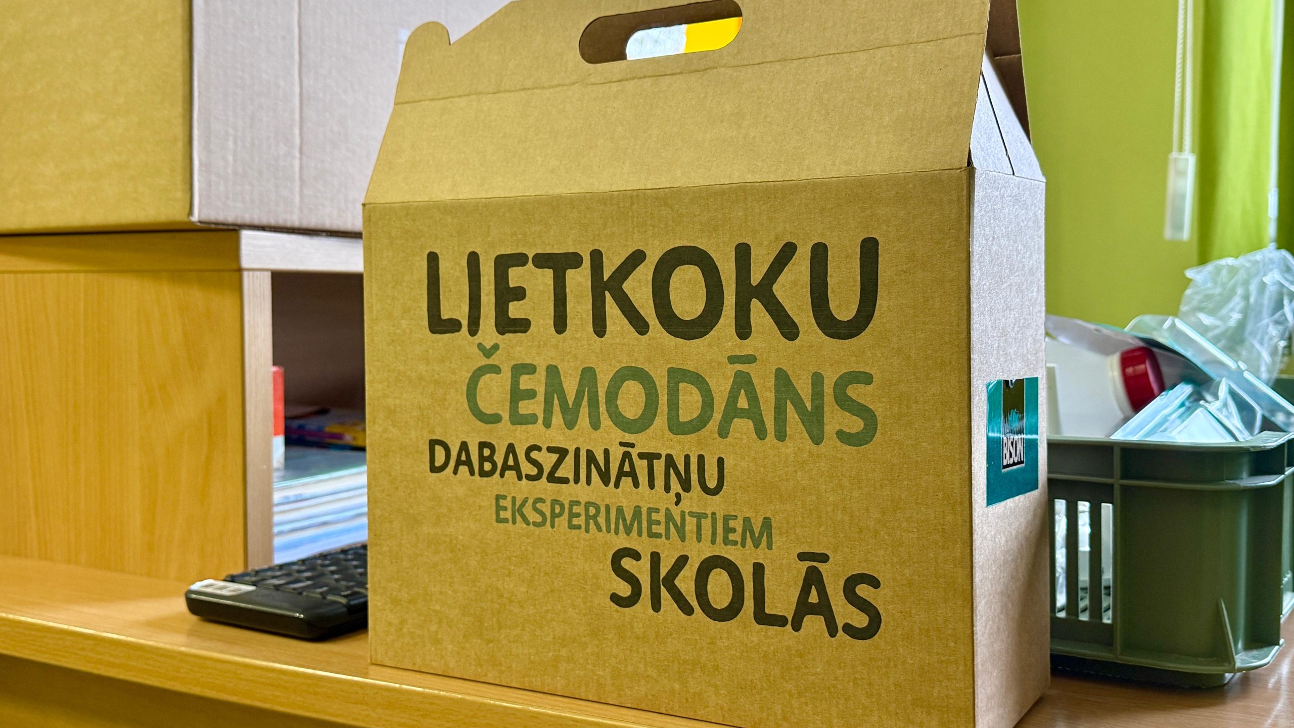Brūna kartona kaste ar lielu uzrakstu: "LIETKOKU ČEMODĀNS DABASZINĀTŅU EKSPERIMENTIEM SKOLĀS"