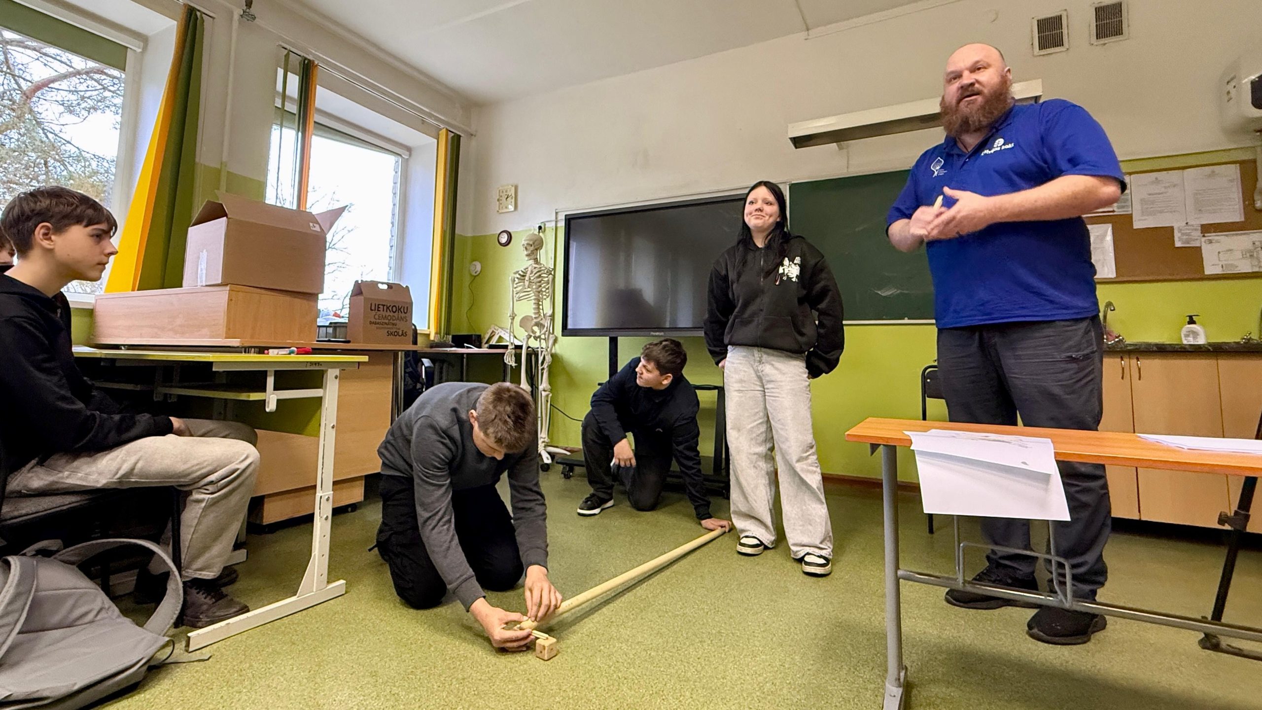 Skolēni mācību stundā veic eksperimentu ar koka klučiem un koka stieni, ko uzrauga 