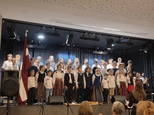 Bērnu un jauniešu kolektīvs tautas tērpos un svētku apģērbā dzied Latvijas valsts svētku koncertā, blakus Latvijas karogs.