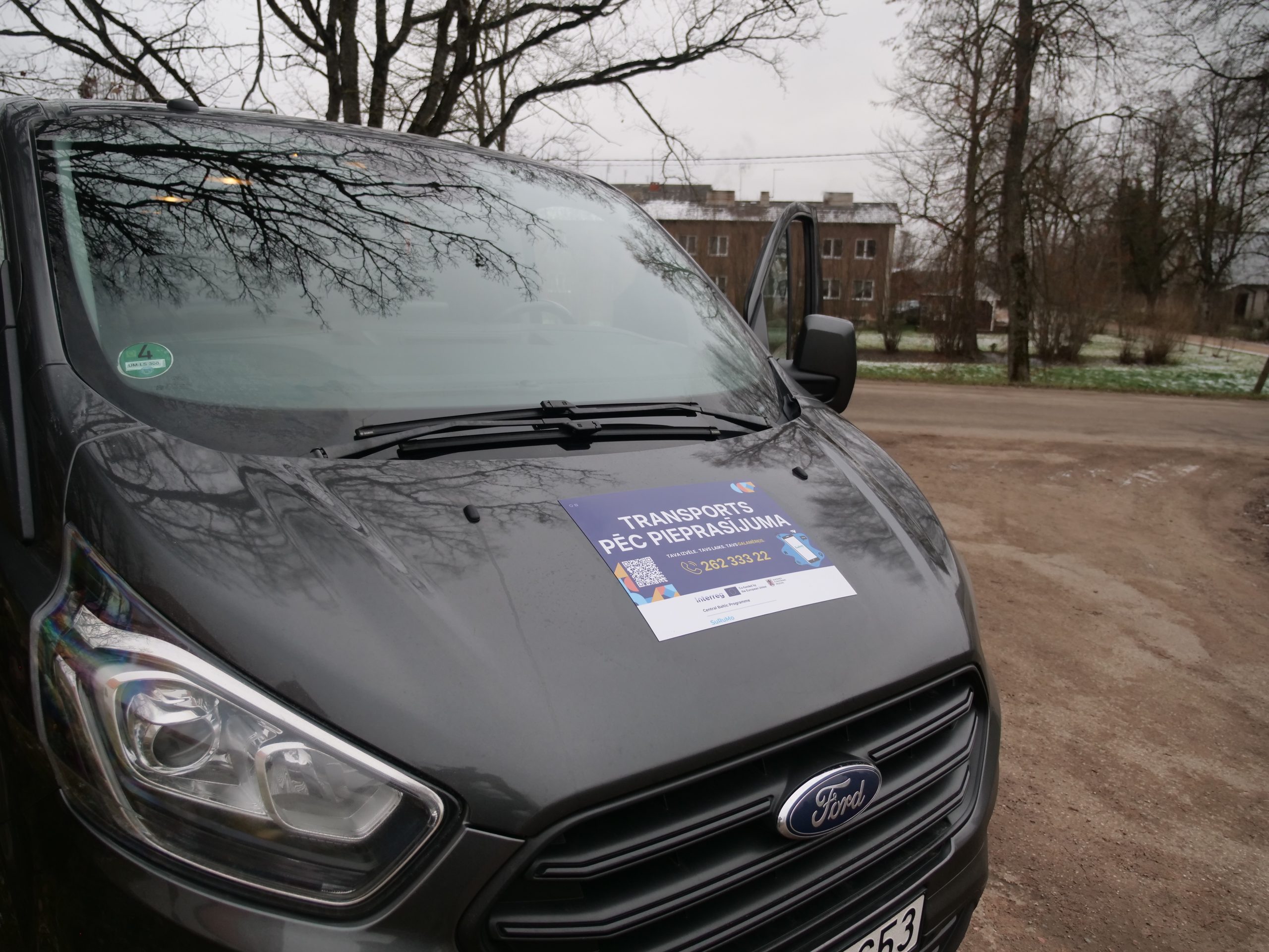 Transports pēc pieprasījuma mikroautobuss ar informatīvu uzlīmi uz motora pārsega.