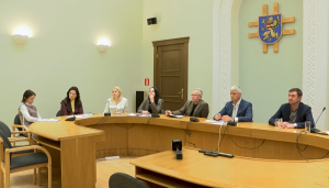 Pašvaldības sēde vai sanāksme pie ovāla galda ar vairākiem dalībniekiem, kuri klausās un pieraksta, fonā redzams pilsētas ģerbonis.