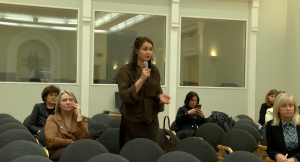 Sieviete ar mikrofonu runā sanāksmē vai publiskā diskusijā, sēžu zālē ar vairākiem klausītājiem fonā.