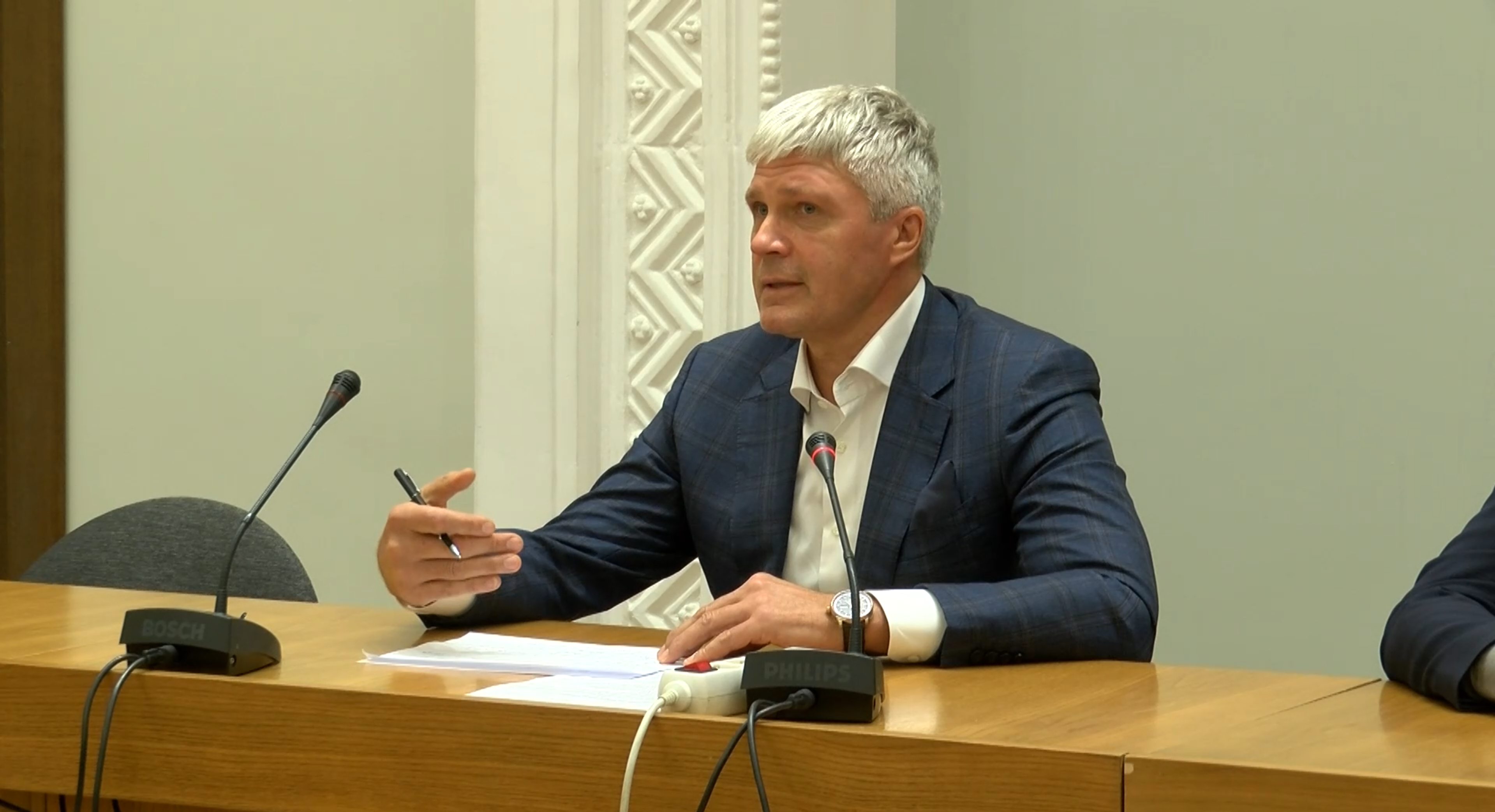 Aleksandrs Bartaševičs runā sēdes laikā, sēžot pie mikrofoniem un ar pildspalvu rokā.