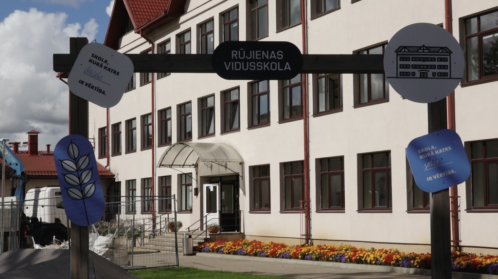 Rūjienas vidusskolas ēka ar ieeju un uzrakstu “Skola, kurā katrs solis ir vērtība”