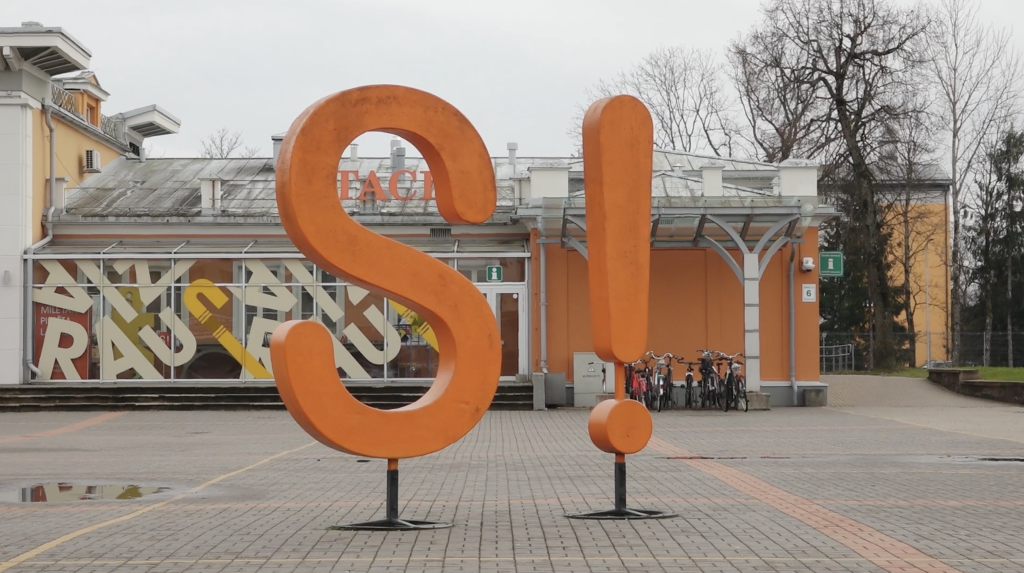 Liels, oranžs S burts un izsaukuma zīme Siguldas pilsētvidē.
