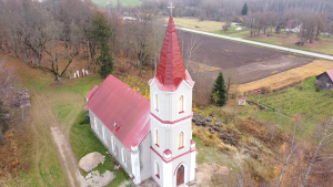 Seces evaņģēliski luteriskā baznīca ar sarkanu torni, kas redzama no drona lidojuma, fonā lauku ainava un Latvijas karogs.