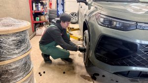 Autoservisa darbinieks nomaina riepas tumšpelēkai automašīnai, kas ir pacelta uz pacēlāja.