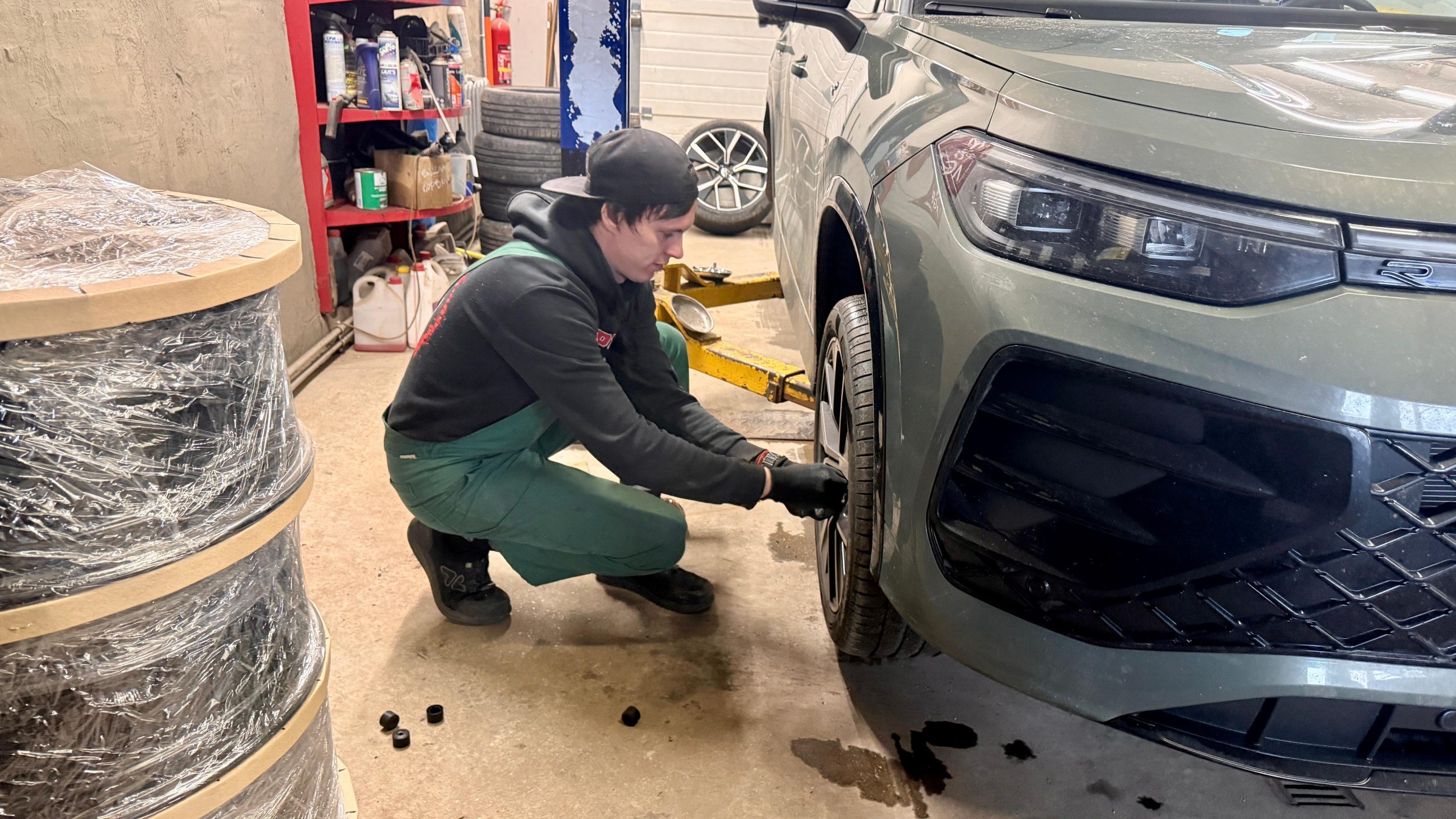 Autoservisa darbinieks nomaina riepas tumšpelēkai automašīnai, kas ir pacelta uz pacēlāja.