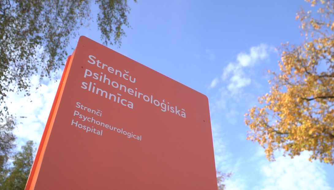Strenču psihoneiroloģiskās slimnīcas informatīvā zīme pie ieejas ēkā saulainā dienā.