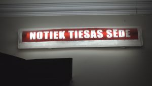 Spilgti sarkans uzraksts uz gaismas paneļa "NOTIEK TIESAS SEDE" (Notiek tiesas sēde).