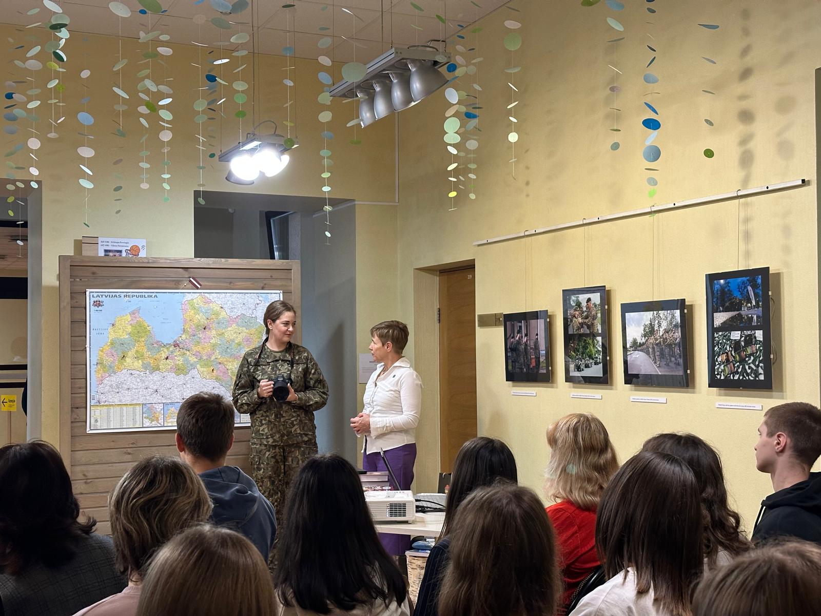 Sieviete militārā formā un pasākuma vadītāja runā skatītāju priekšā pie Latvijas kartes, telpā ar fotogrāfiju izstādi uz sienas.
