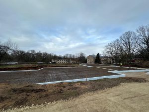 Būvlaukums ar ieklātu armējumu un pamatu kontūrām, fonā redzamas daudzdzīvokļu ēkas un strādnieks drošības vestē.