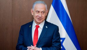 Izraēlas premjerministrs Benjamins Netanjahu stāv pie Izraēlas karoga, uzvalkā ar sarkanu kaklasaiti.