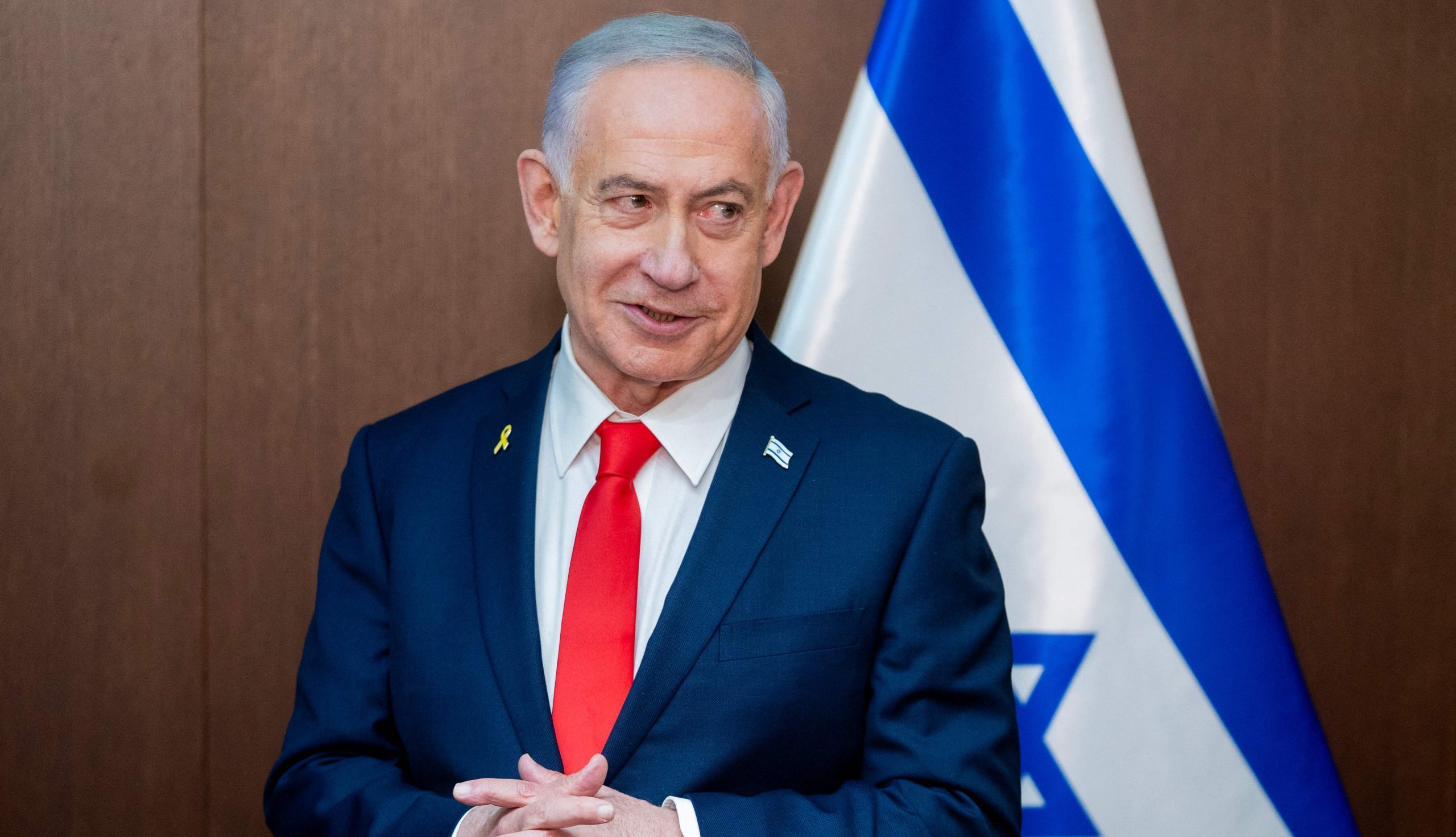 Izraēlas premjerministrs Benjamins Netanjahu stāv pie Izraēlas karoga, uzvalkā ar sarkanu kaklasaiti.