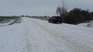 Ziemā piesalis grants ceļš Bauskas novadā ar ceļa malā novietotu automašīnu.
