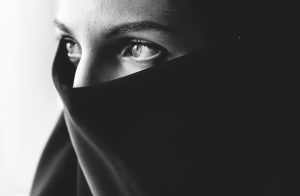 Melnbalts, ļoti tuvs plāns, kurā redzamas sievietes acis un piere, seja daļēji aizsegta ar tumšu audumu, kas atgādina nikābu vai hidžābu.